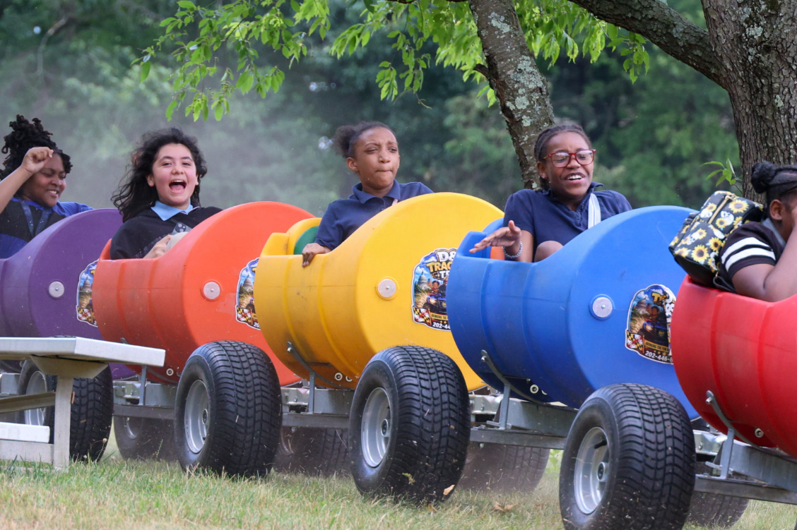 Girls laughing and cheering in colorful barrel train cars under the trees