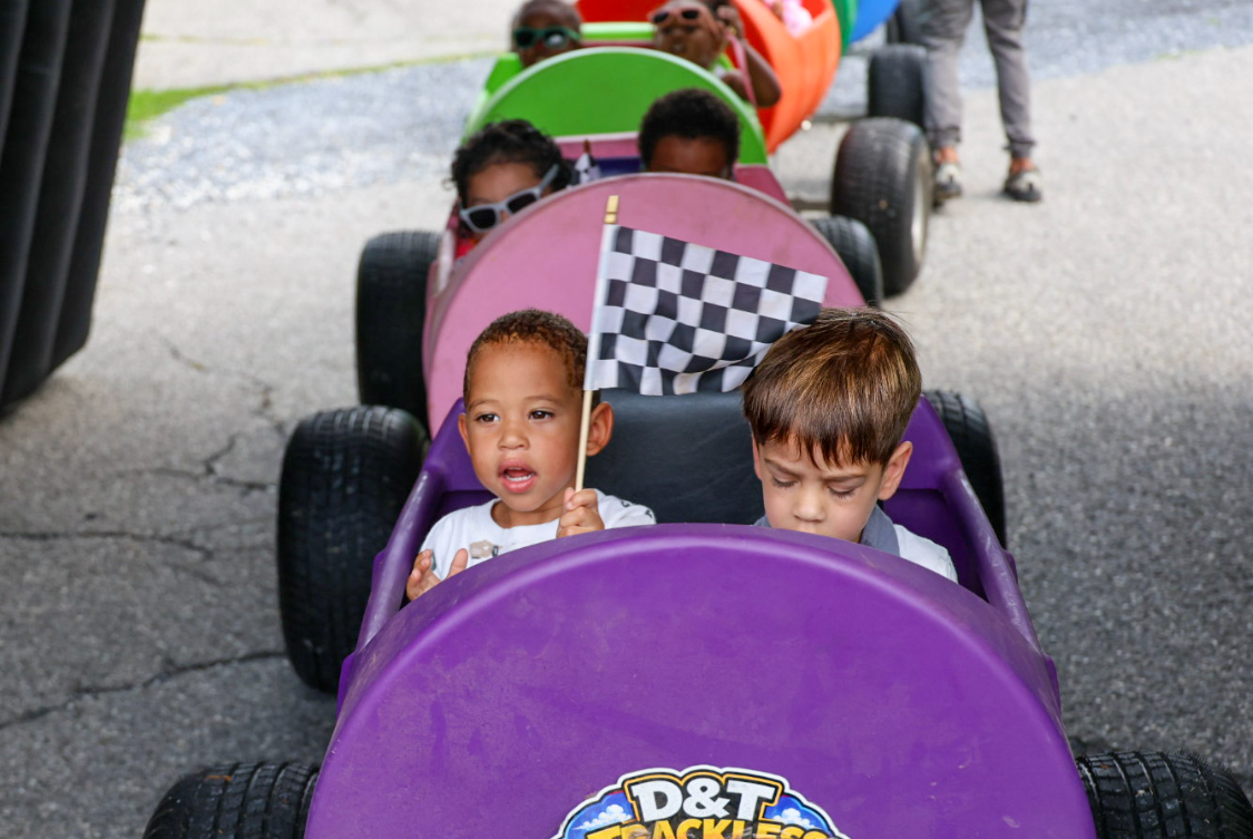 Toddlers riding in the purple D&T Trackless Train car holding a checkered flag